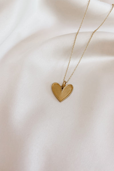 Darling Necklace in gold is shown close up on silk.  It has a beautiful matte finish and organic raised edges on a uniquely shaped large heart pendant.
