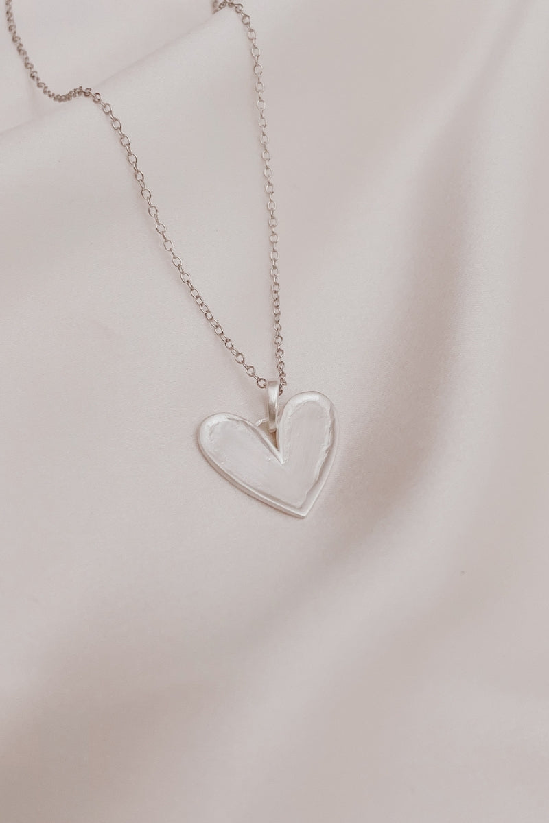 A close up look at the Darling Necklace in sterling silver with its slightly raised, organic shaped edges and soft matte finish.  The unique heart pendant hangs from a Fine Cable chain.