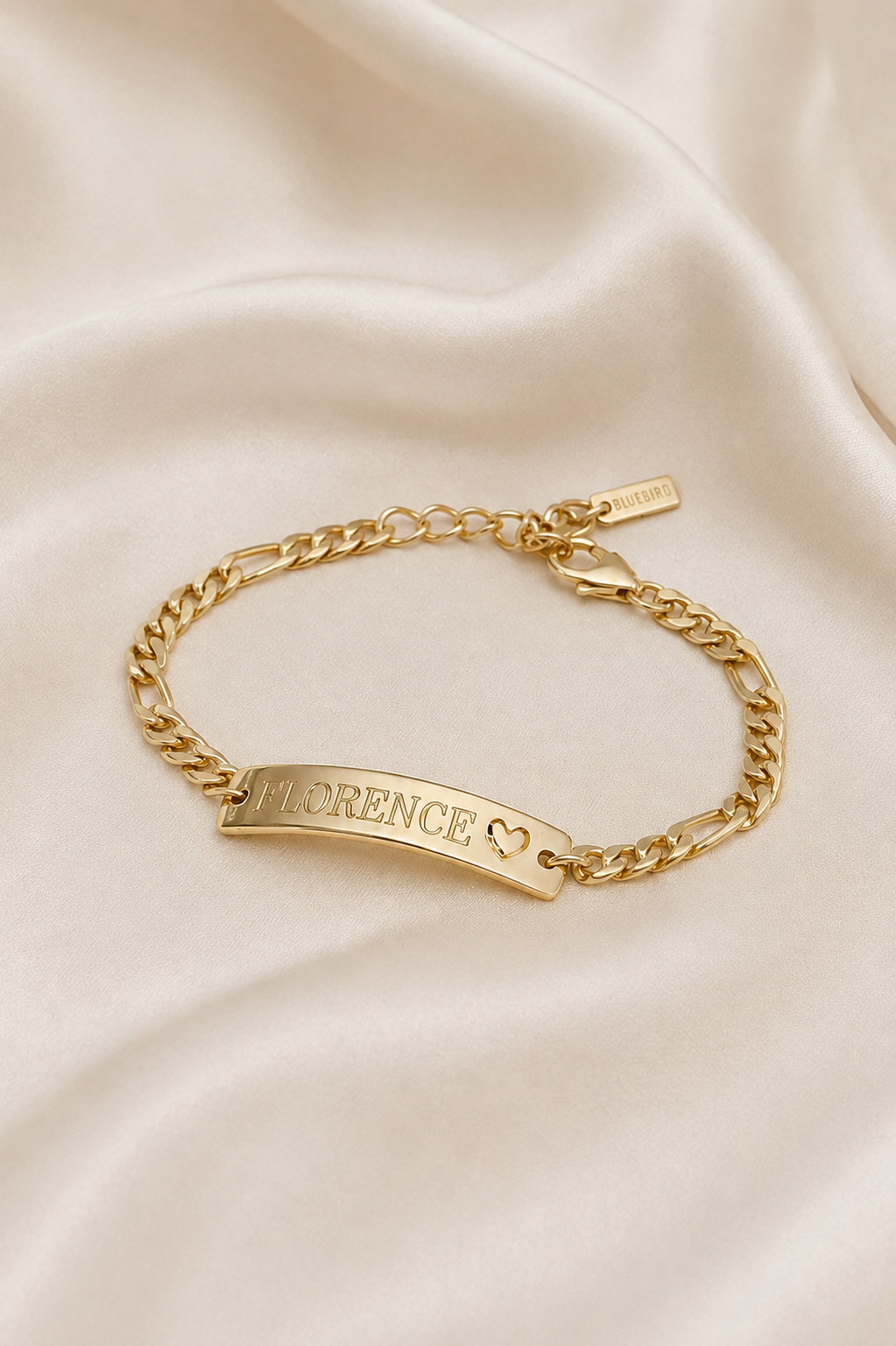 A gold chain bracelet with a curved ID plate engraved with the name 'FLORENCE' and a small heart, displayed on a draped cream satin fabric background.