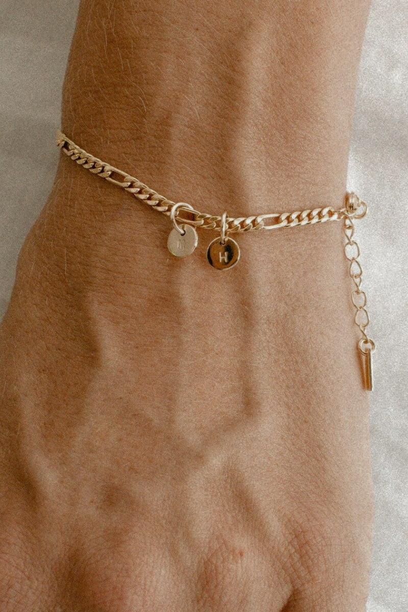 A Figaro Bracelet with two solid gold 6mm initial pendants rests elegantly on a woman's wrist.