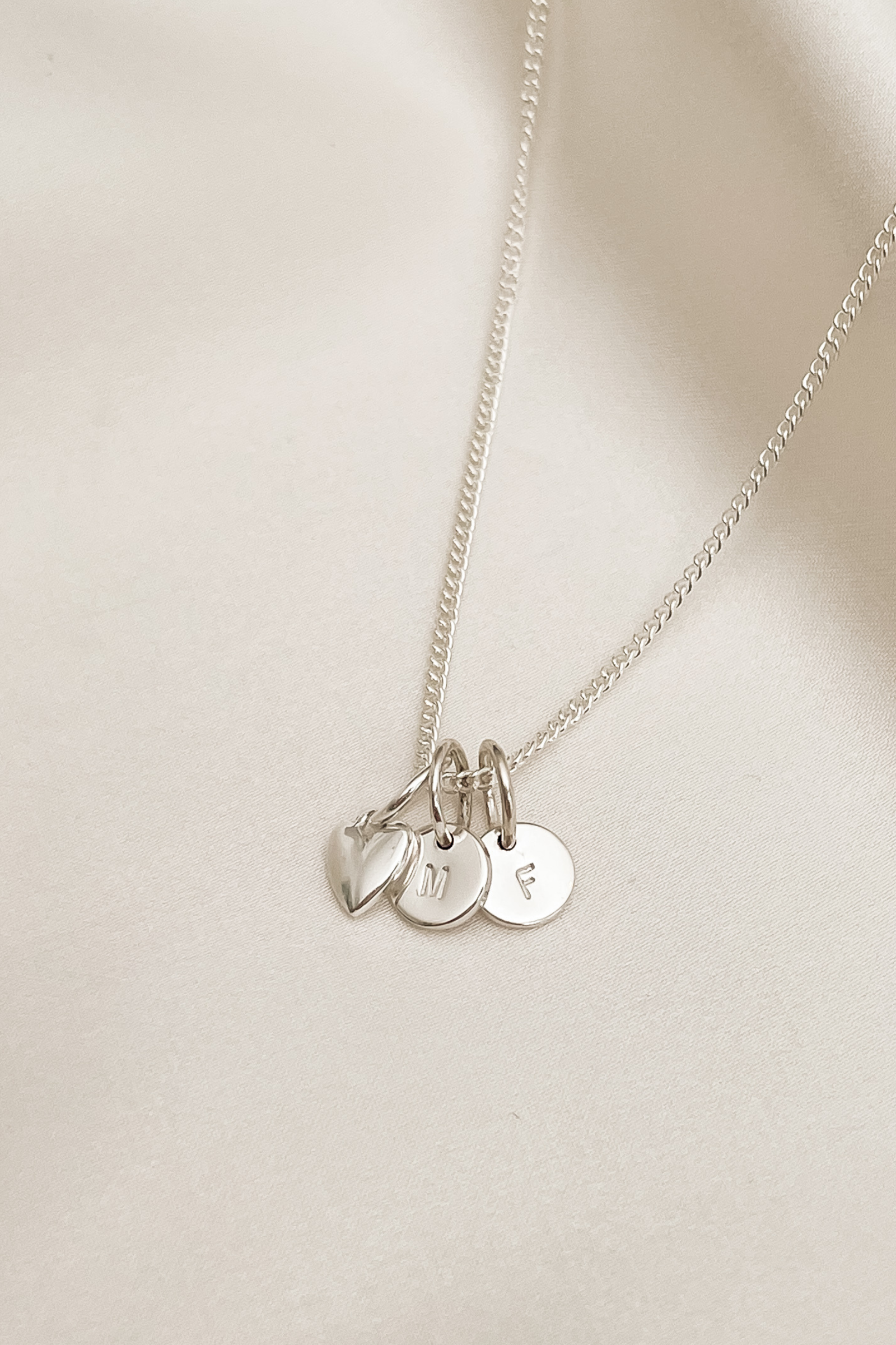 A silver fine curb chain has a mini sweetheart pendant and two initial pendants on it.  