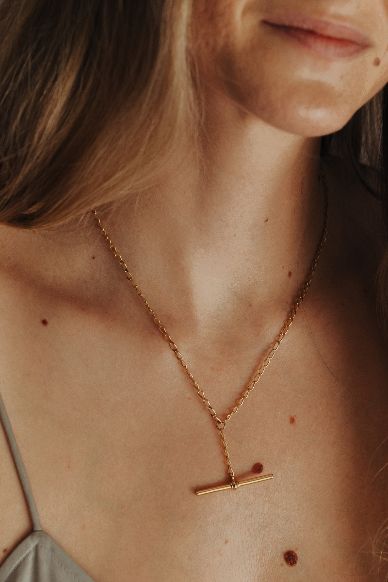 A Gold Fob Necklace also known as a T-Bar Necklace is worn on a woman and features an elegant curb chain with T-Bar drop chain moving freely along the chain.