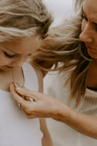 A mother is taking a close look at the engraved 'M' on the front of her daughter's Bluebird Co Jewellery Petite Signature Necklace.