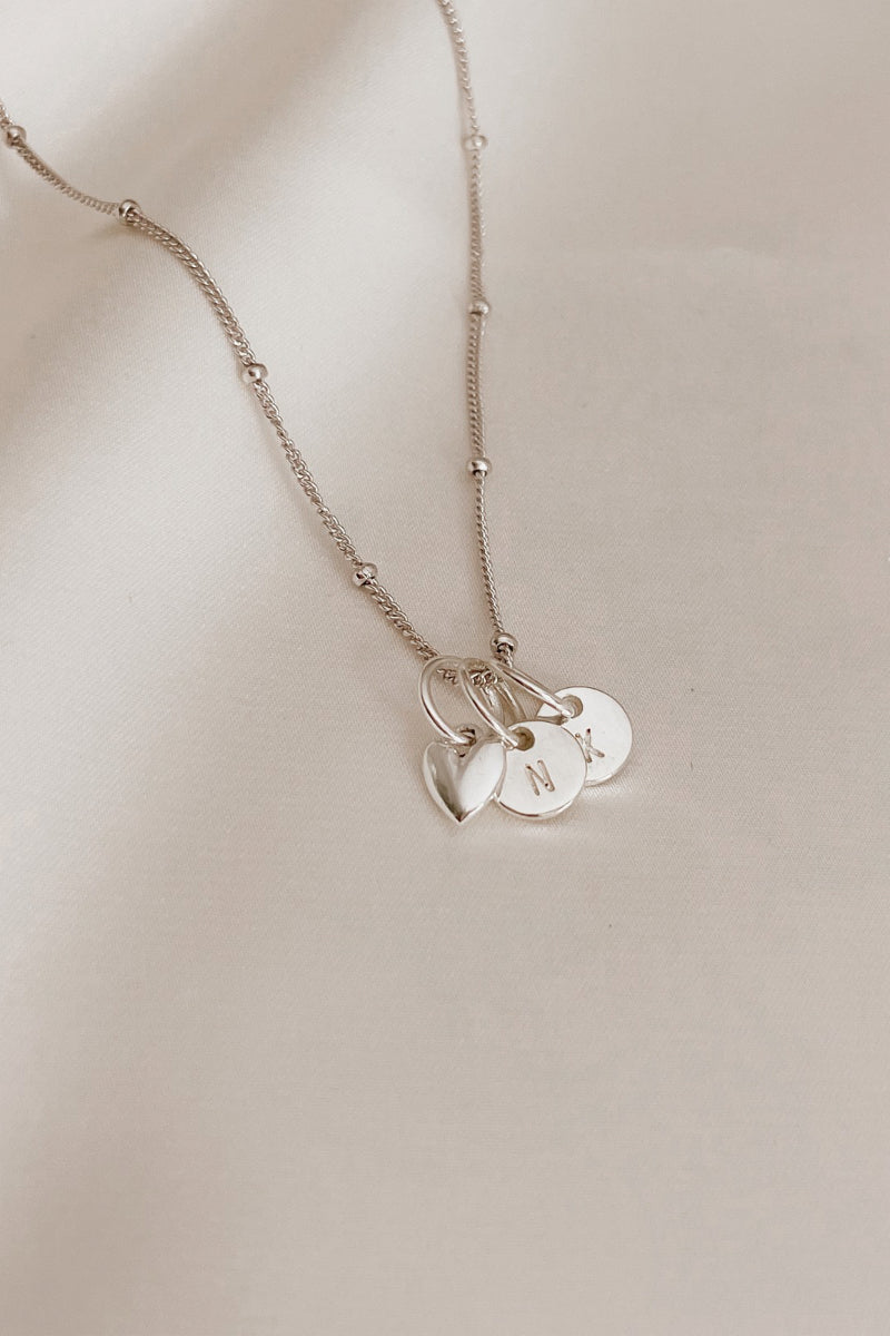 A sterling silver necklace chain with a heart pendant and two petite initial pendants that are round.