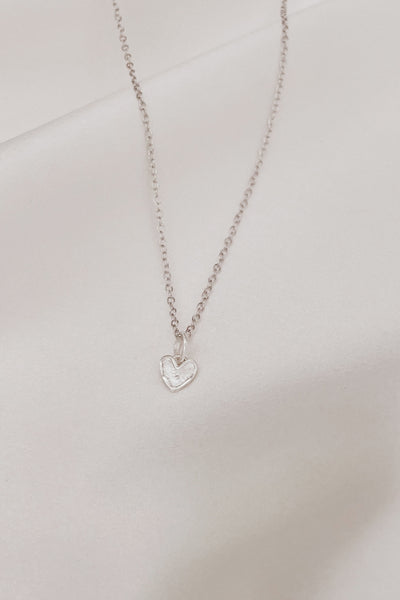 A Petite Darling Necklace is shown on silk with its fine cable chain and uniquely textured surface with raised edges, this petite heart shaped pendant is timeless and stylish and can be personalised by adding Initial Pendants to the chain.