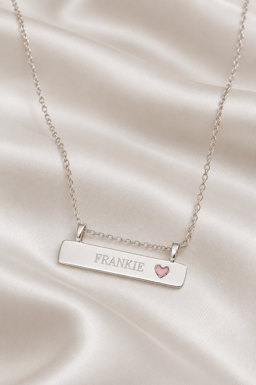 A silver bar necklace with the name 'FRANKIE' engraved on the front and a small pink heart accent, displayed on draped white satin fabric.