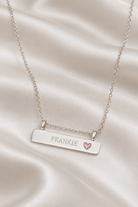 A silver bar necklace with the name 'FRANKIE' engraved on the front and a small pink heart accent, displayed on draped white satin fabric.