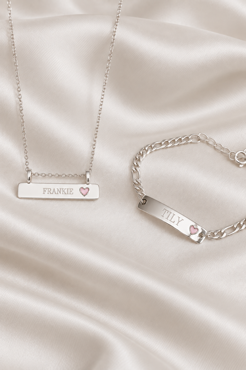 A silver necklace and matching bracelet displayed on draped satin fabric. The rectangular nameplates are engraved with the names 'FRANKIE' and 'TILY' respectively, and each has a small pink heart accent.
