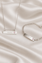 A silver necklace and matching bracelet displayed on draped satin fabric. The rectangular nameplates are engraved with the names 'FRANKIE' and 'TILY' respectively, and each has a small pink heart accent.