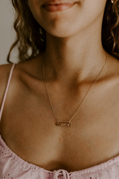 A teenage girl is wearing a soft pink top with her Darling Bar Necklace that has personalised engraving on it.  She has her name engraved on the front of the bar pendant and wears the 45+5cm length.