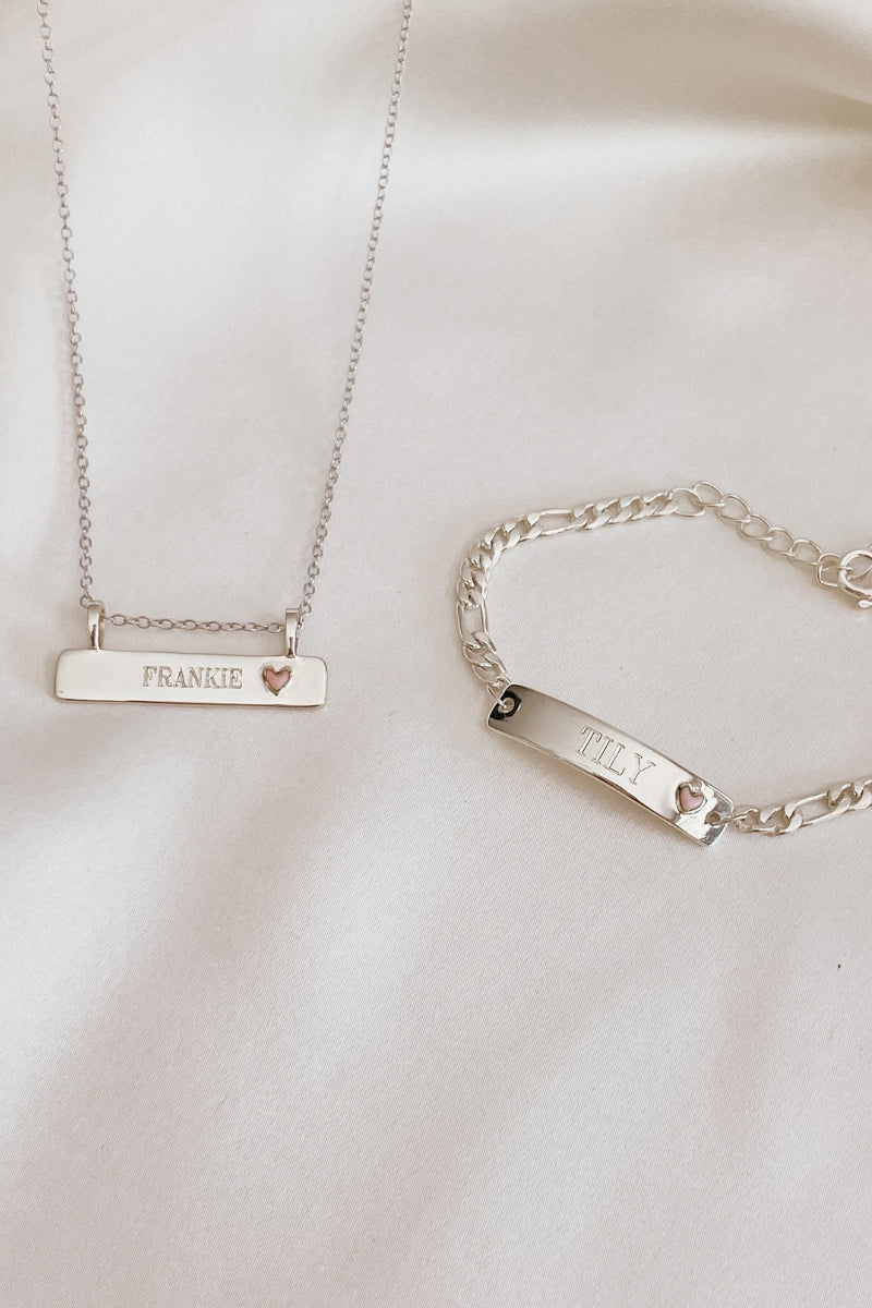 A Pink Darling Bra Necklace is engraved with a child's name on the pendant and it is sitting alongside the Pink Darling Bracelet which is also engraved with a child's name on the bar pendant.