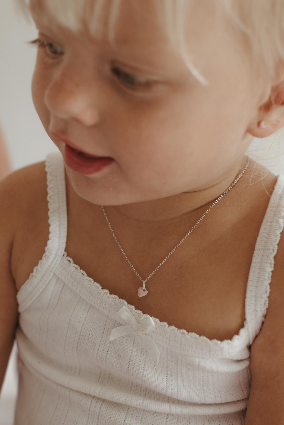 A young girl wears a soft pink coloured heart pendant necklace on a solid sterling silver chain.  It can be personalised with an initial pendant