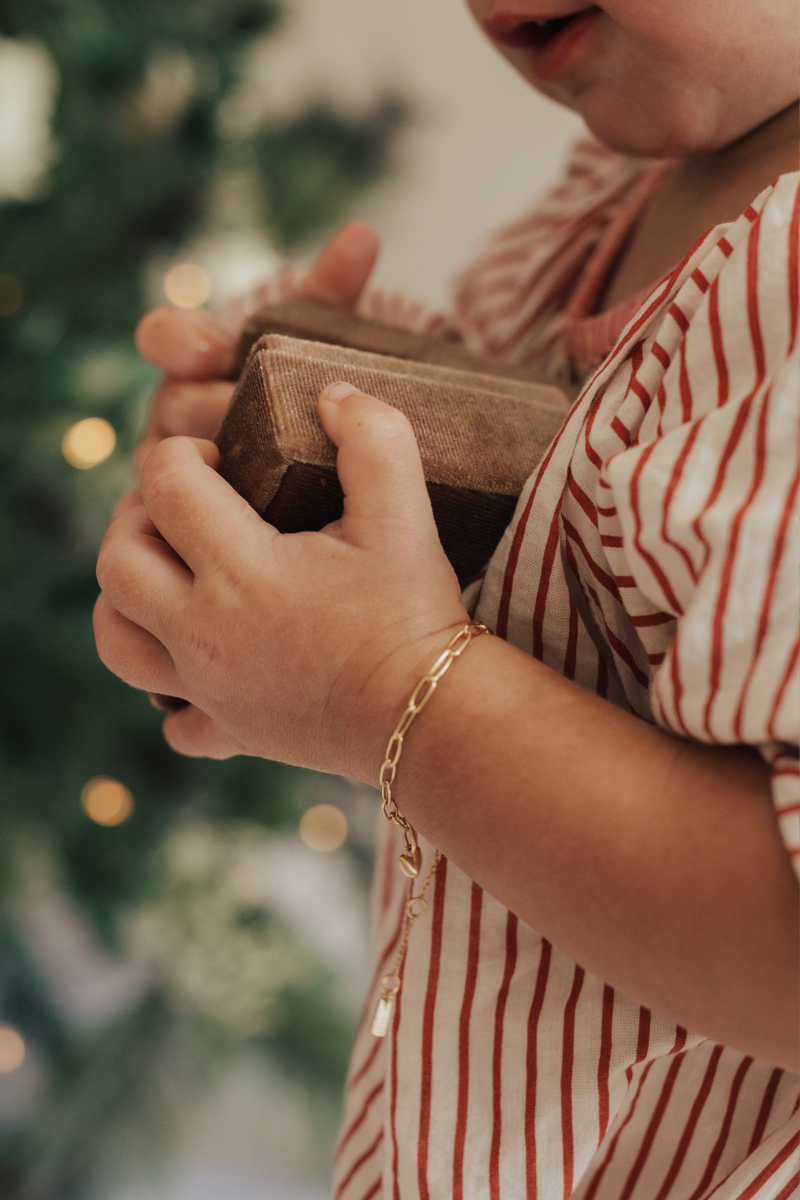 A baby wearing a gold Posie Bracelet with a Mini Sweetheart pendant, holding a velvet box captured in natural light to highlight its delicate craftsmanship by Bluebird Jewellery.