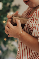 A baby wearing a gold Posie Bracelet with a Mini Sweetheart pendant, holding a velvet box captured in natural light to highlight its delicate craftsmanship.