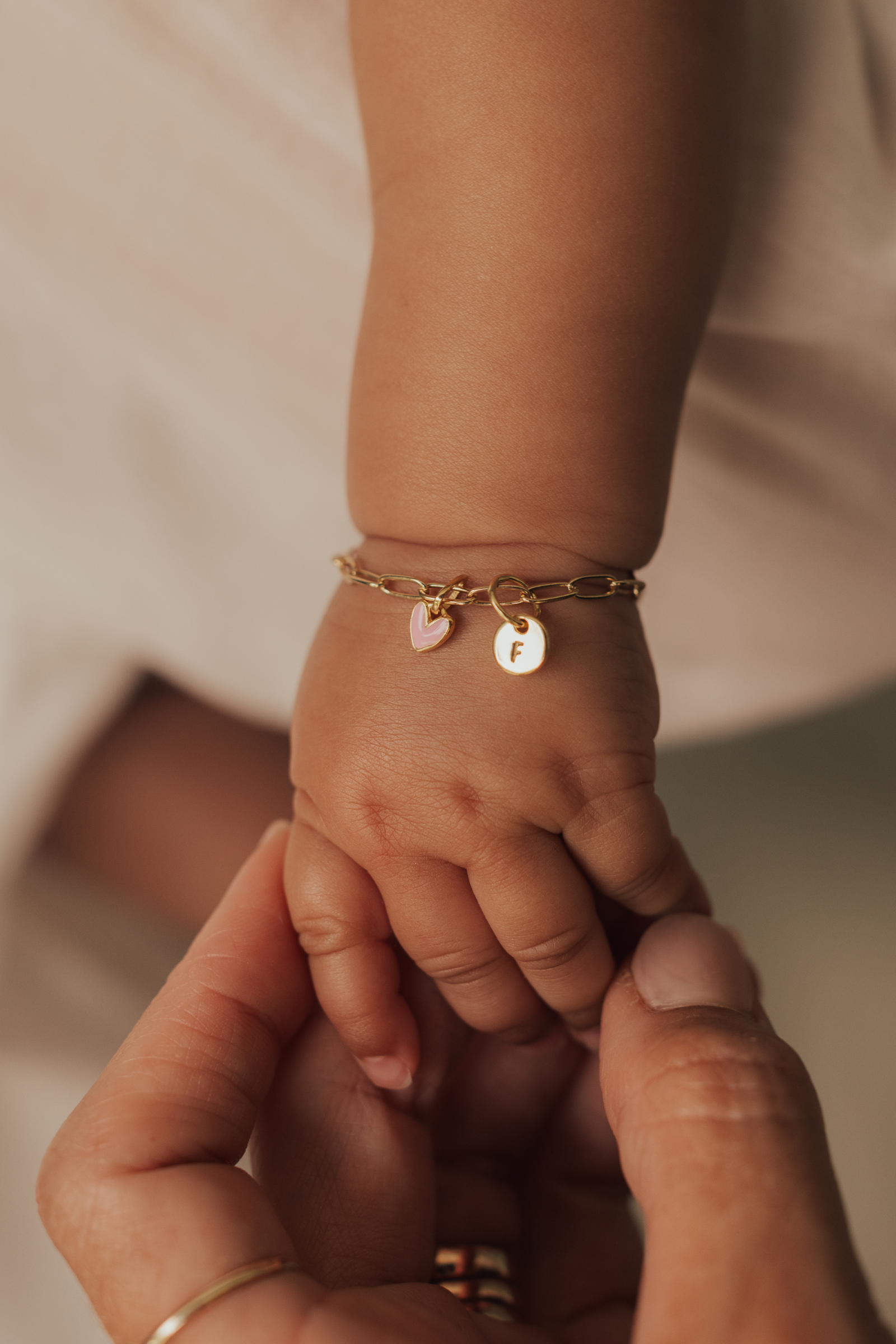 A mother is holding a baby's hand and the baby is wearing a gold baby bracelet with pink heart pendant and petite round initial pendant. Bluebird Jewellery.
