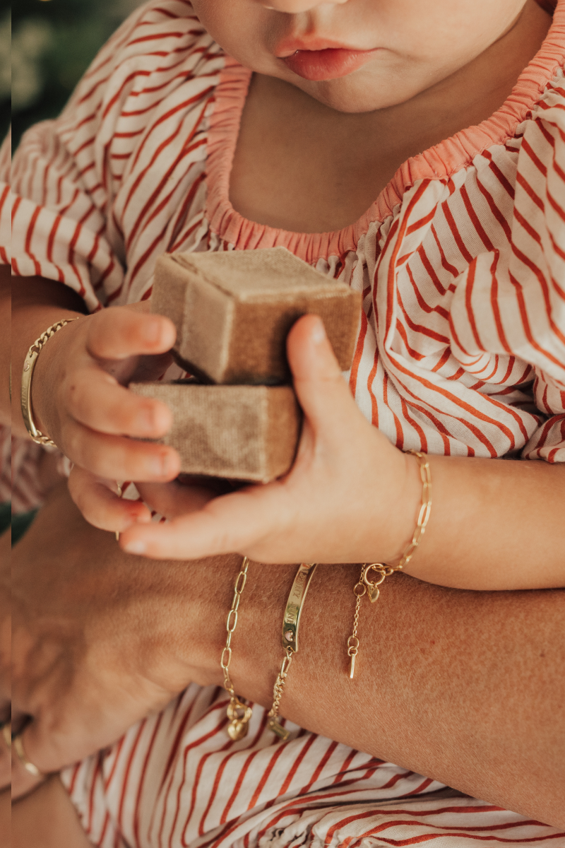 A close-up of a mother and child’s hands featuring gold link chain and personalised bar pendant bracelets, symbolising connection and timeless craftsmanship by Bluebird Jewellery.