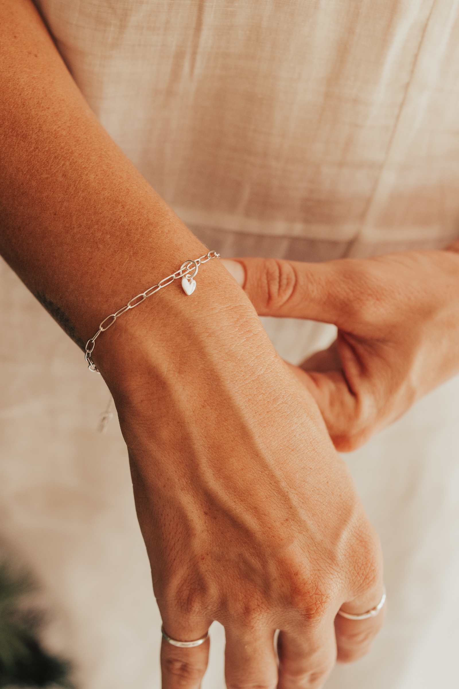 A close-up of a hand wearing a Posie Bracelet with a heart charm, complemented by delicate silver rings for a refined modern finish by Bluebird Jewellery 