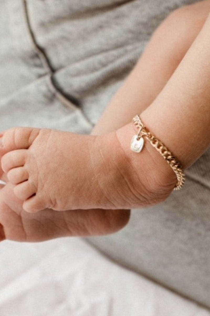 A close-up of a baby’s ankle wearing a gold Cuban Link Chain Bracelet with a solid rectangular pendant, crafted as a delicate keepsake piece.