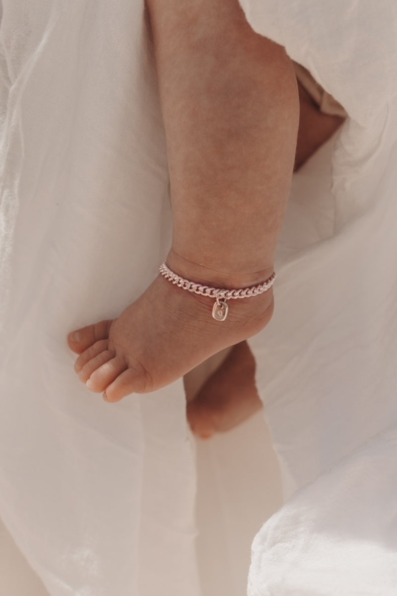 A close-up of a baby’s ankle wearing a rose gold Cuban Link Chain Bracelet with a solid gold rectangular pendant, crafted as a delicate keepsake piece.