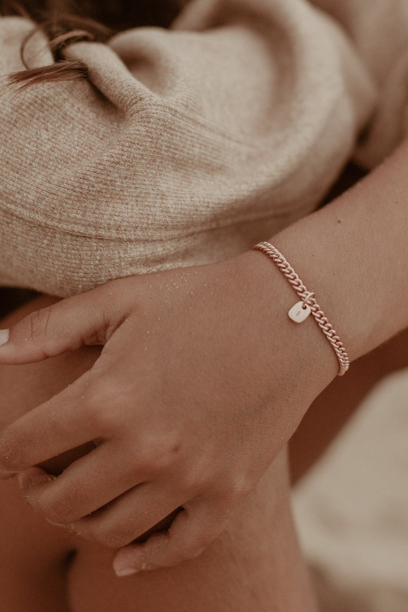 A refined image of a woman’s wrist wearing a rose gold Cuban Link Chain and solid gold rectangular pendant, photographed in natural light by the seaside by Bluebird Jewellery.