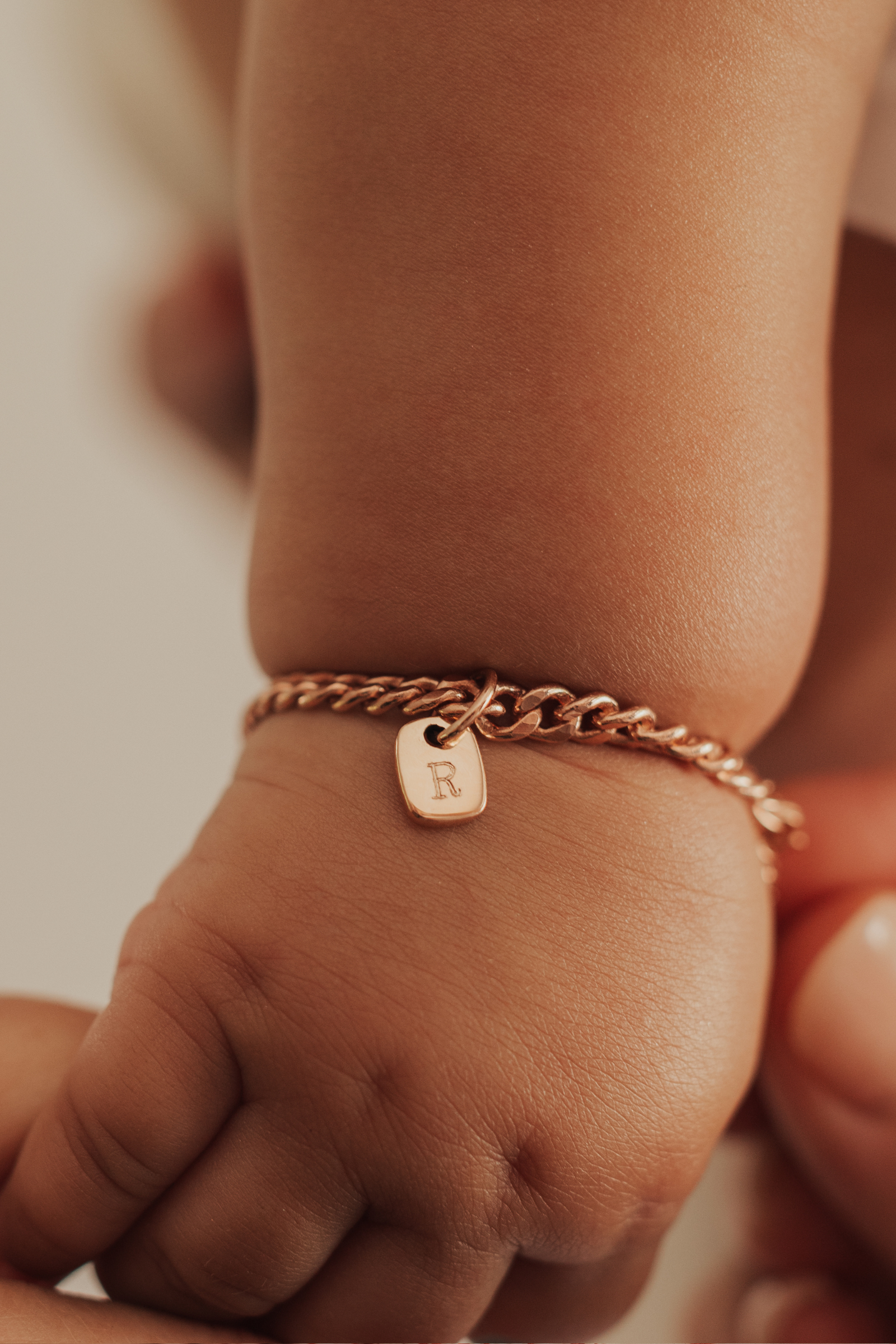 A close-up of a baby’s wrist wearing a rosegold Cuban Link Chain Bracelet with a soild gold rectangular pendant, crafted as a delicate keepsake piece by Bluebird Jewellery.