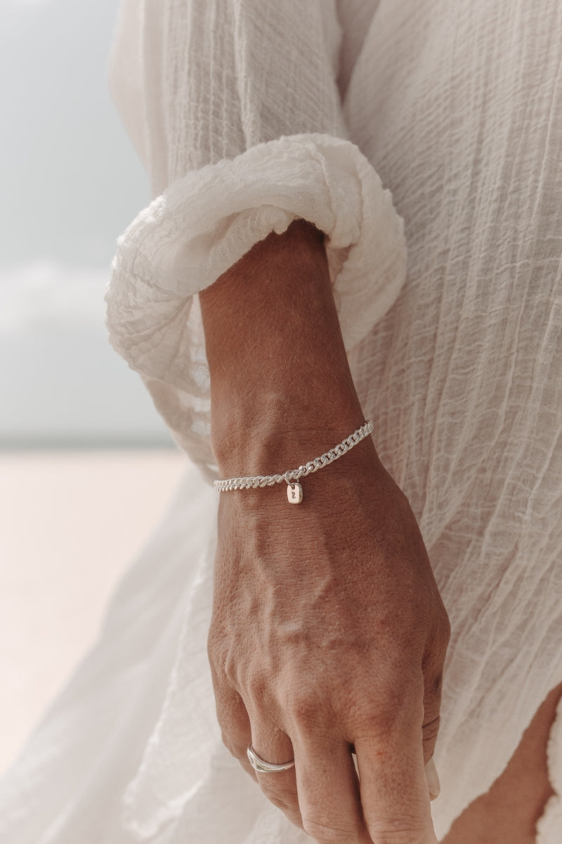 A close-up photo of a woman’s wrist at the beach featuring a Cuban Link Chain with a minimalist rectangular pendant and a matching signet ring, styled in fine jewellery fashion by Bluebird Co