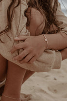An image of a woman’s wrist adorned with a rose gold Cuban Link Chain and soild gold rectangular pendant, photographed by the ocean for a relaxed, elegant look.