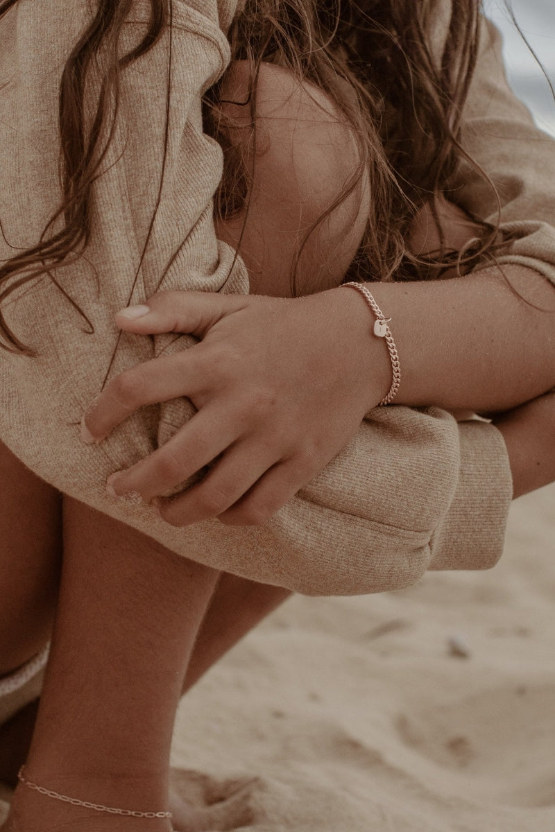 An image of a woman’s wrist adorned with a rose gold Cuban Link Chain and soild gold rectangular pendant, photographed by the ocean for a relaxed, elegant look.