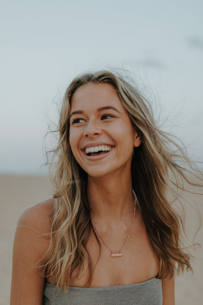 Rose gold Signature Bar Necklace shown on model laughing at the beach. Personal and stylish jewellery for everyday wear. 