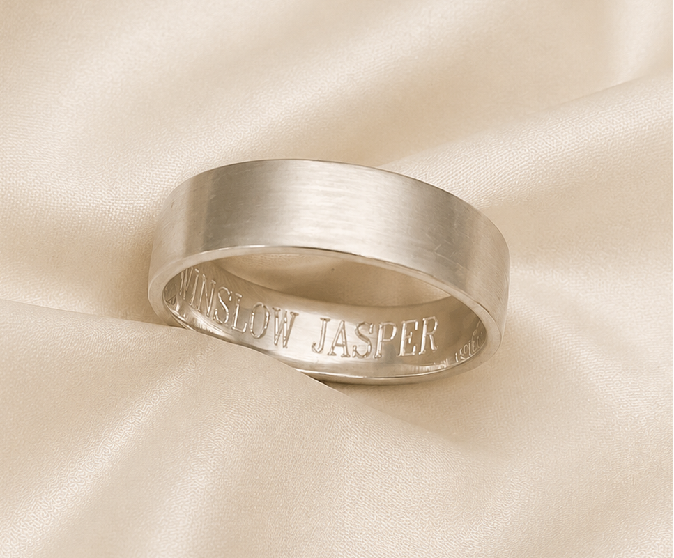 A brushed silver band ring with 'JASPER' engraved on the inside, resting on draped cream satin fabric.