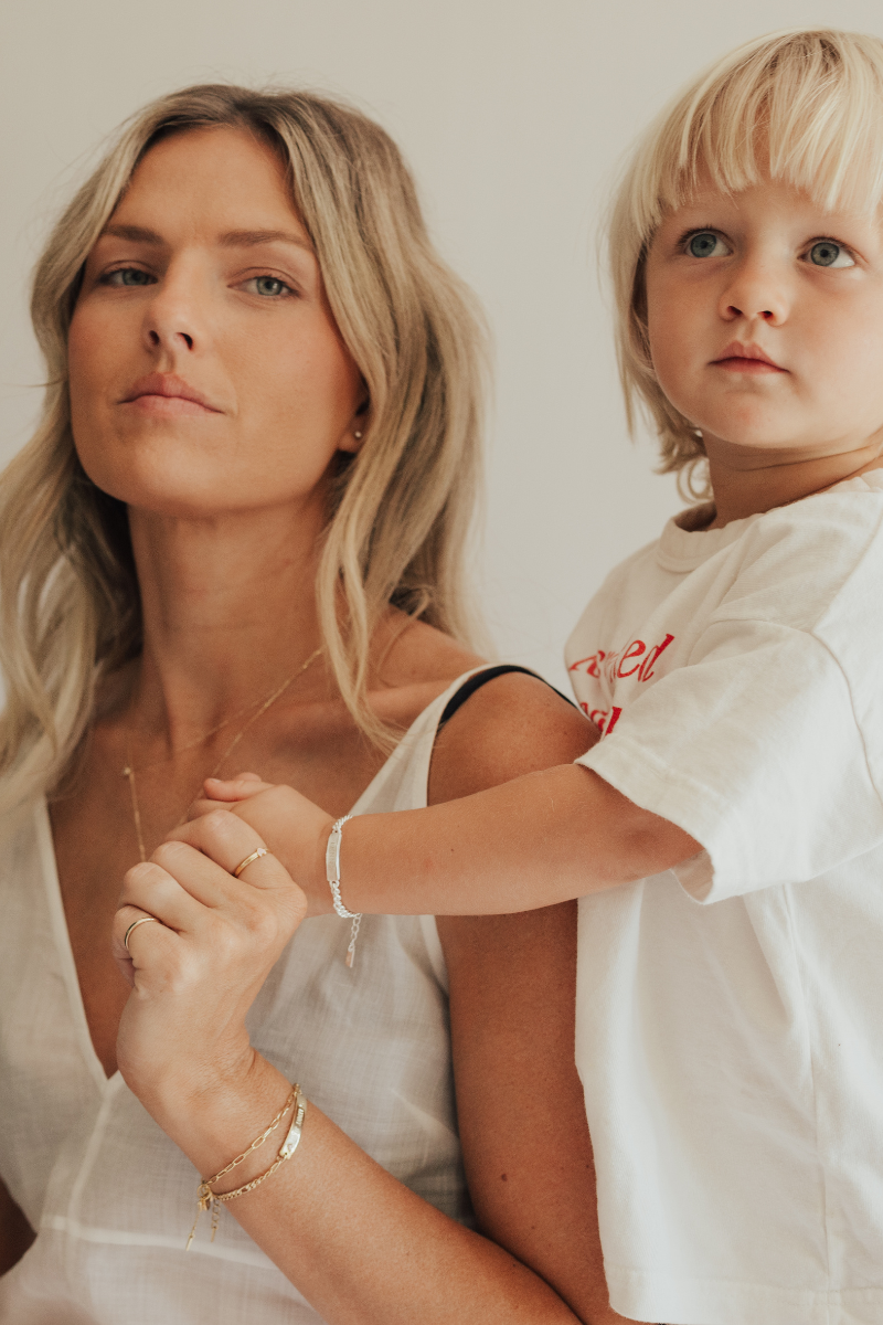 A mother is holding hands with her son.  He is wearing a baby sized Signature Bracelet by Bluebird Jewellery that has personalised engraving on the front of the bracelet.