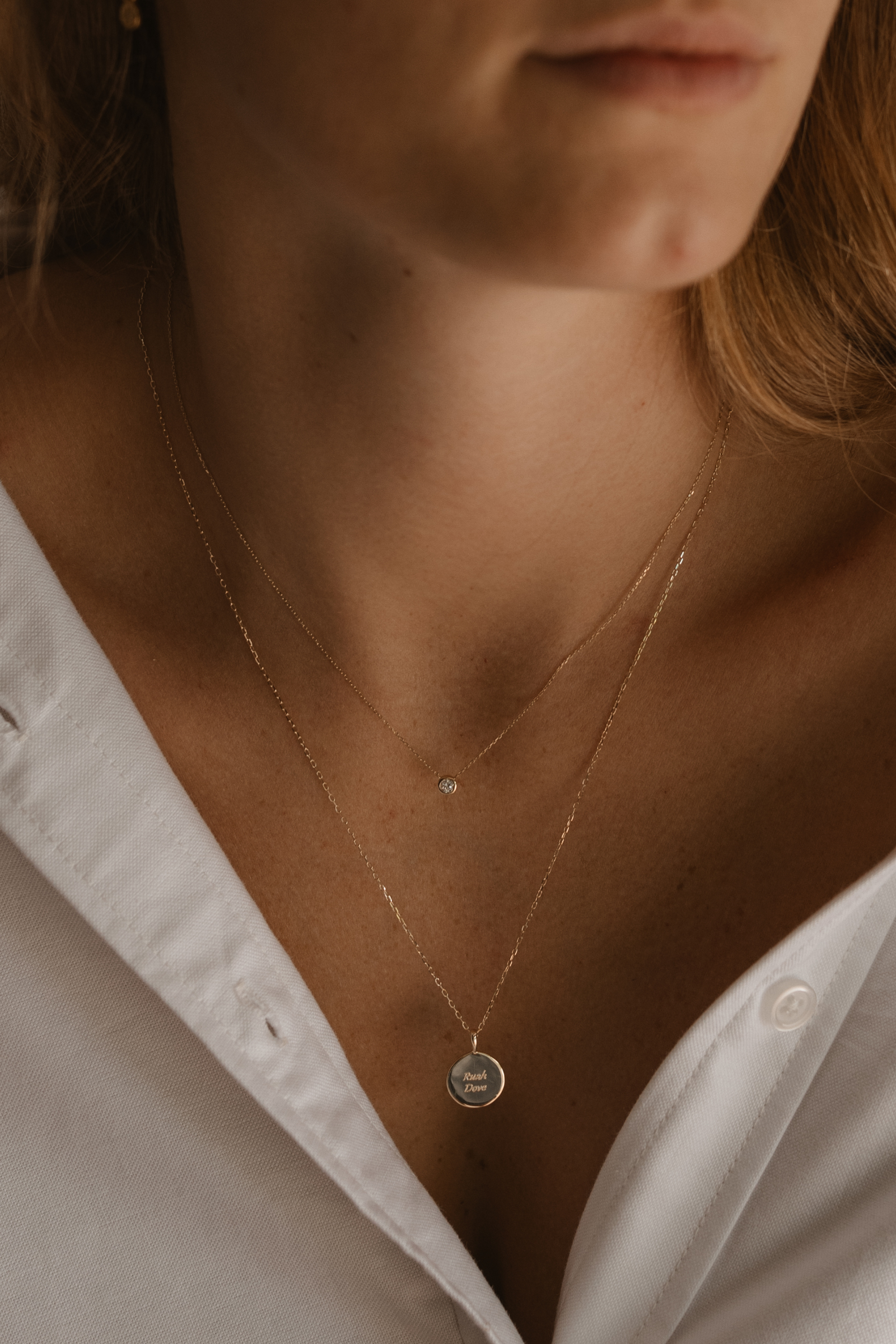 A woman wears a fine diamond necklace layered with a solid gold chain with an engraved pendant on it.