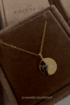 A polished Soleil Necklace in gold with two children's names engraved on the front of it. It is shown in Bluebird Co's signature brown velvet box.