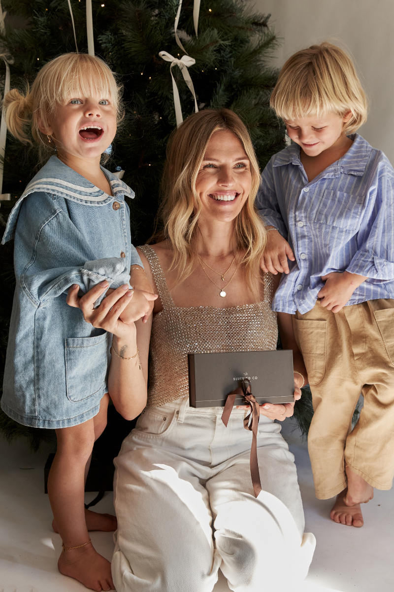 A woman kneels in front of a Christmas tree with two children, holding a dark gray rectangular Bluebird Co gift box with a brown ribbon. The woman wears a sparkly beige top and cream pants, while the children wear casual blue and striped shirts with beige pants.