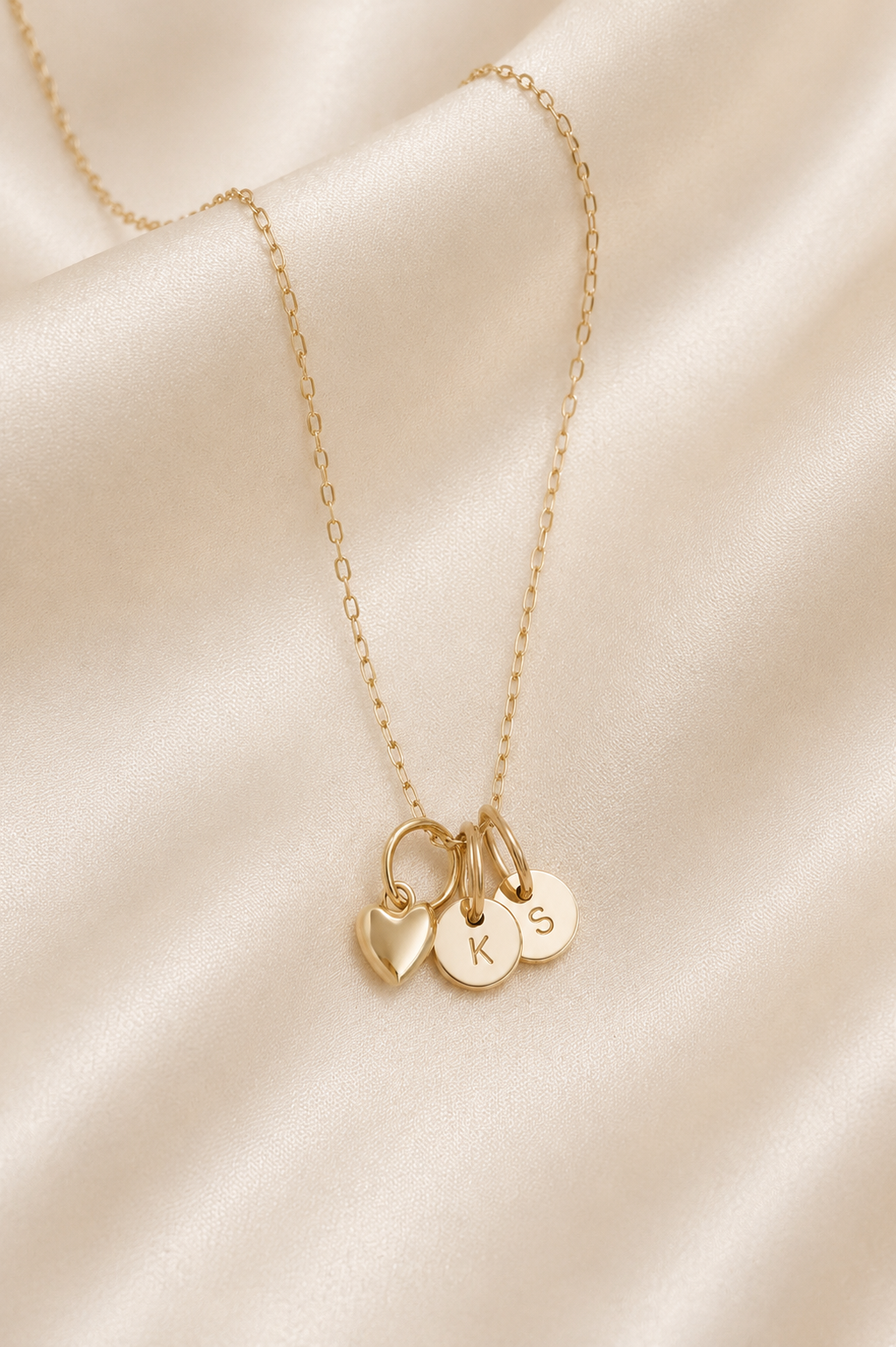 A delicate gold chain necklace with three small hanging charms: a heart, a round initial plate engraved with 'K', and a round initial plate engraved with 'S', displayed on draped champagne satin fabric.
