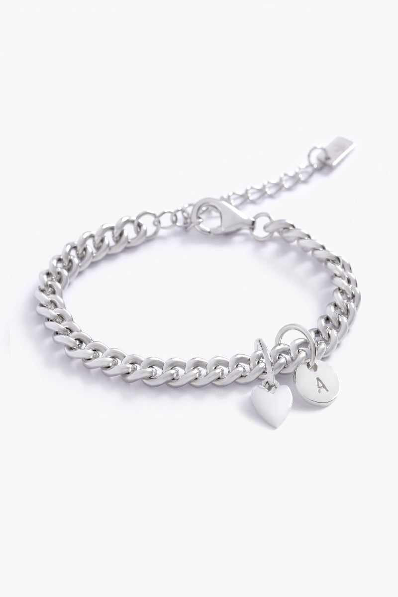 A silver Sweetheart Cuban Link Chain Bracelet adorned with a heart charm and engraved initial pendant, designed as a timeless personalised keepsake by Bluebird Jewellery