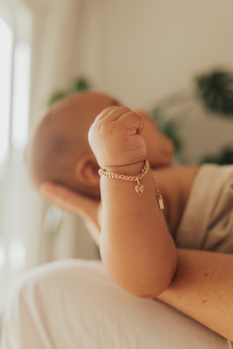 A close up image of a baby wrist wearing a Sweetheart Cuban Link Chain and polished heart pendant, designed as a timeless keepsake piece by Bluebird Jewellery