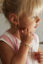 A close-up lifestyle shot of a young girl with blonde hair, wearing a delicate gold chain bracelet with a small heart charm and a matching gold hoop earring, wearing a pink and white striped top.