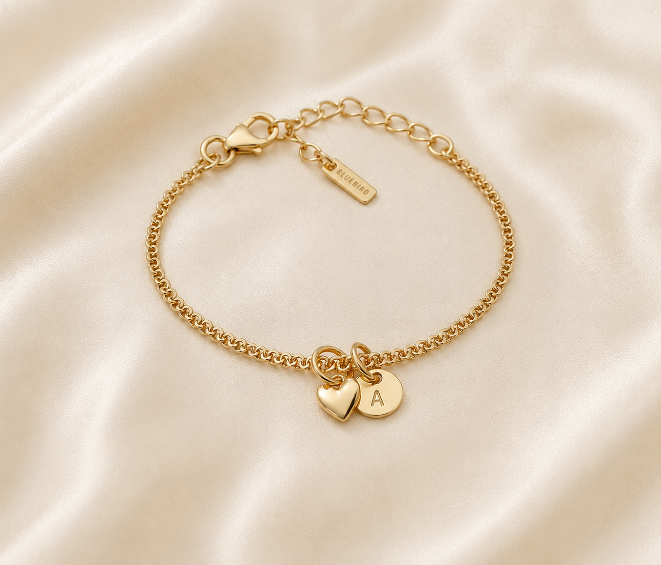 A delicate gold chain bracelet with a lobster clasp, adjustable extender chain, and two small heart charms, one engraved with the letter 'A', displayed on a cream satin fabric background.