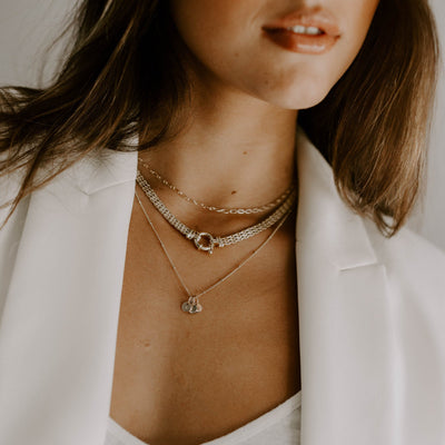 A woman is wearing crisp white singlet and blazer and has three layered necklaces.  The Link Chain Necklace, Signature Statement Necklace and Gold Petite Personalised Necklace