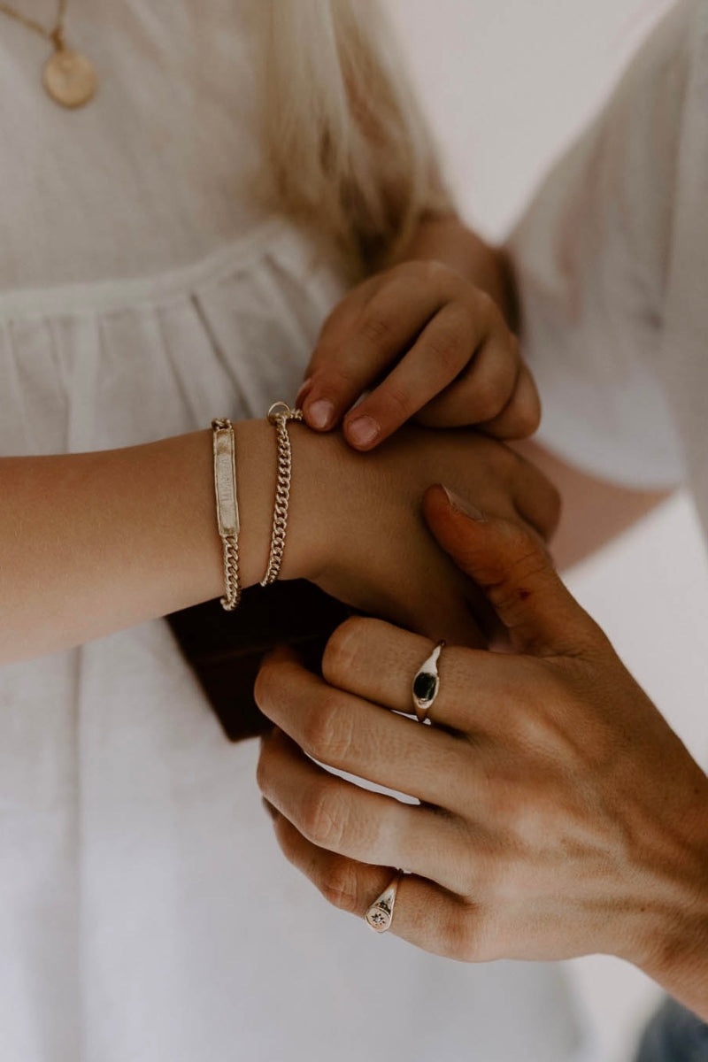 Child wearing engraved gold bracelet while adult helps fasten it, both with gold signet rings, meaningful keepsake jewellery.