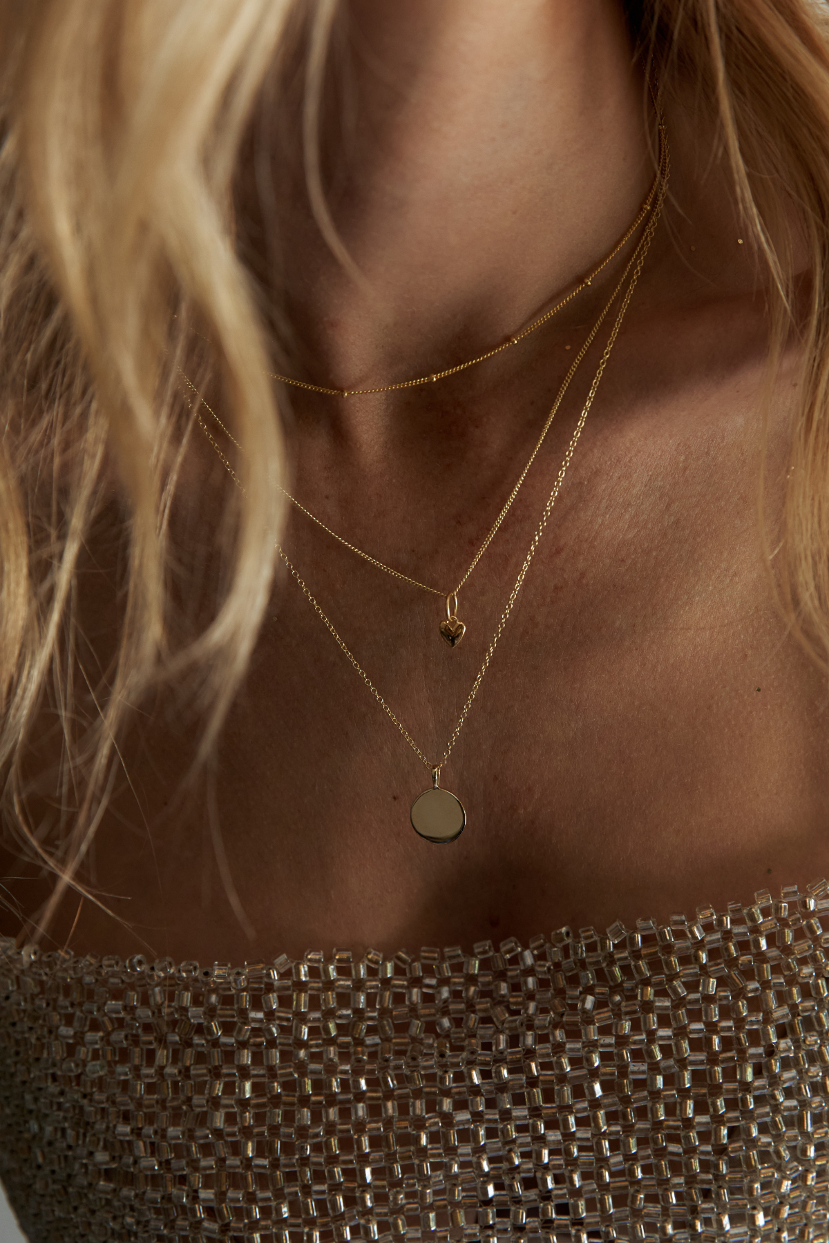 Close-up of a model wearing layered gold necklaces, including a thin chain, a chain with a small heart pendant, and a longer chain with a flat circular disc pendant, paired with a sparkly metallic top.