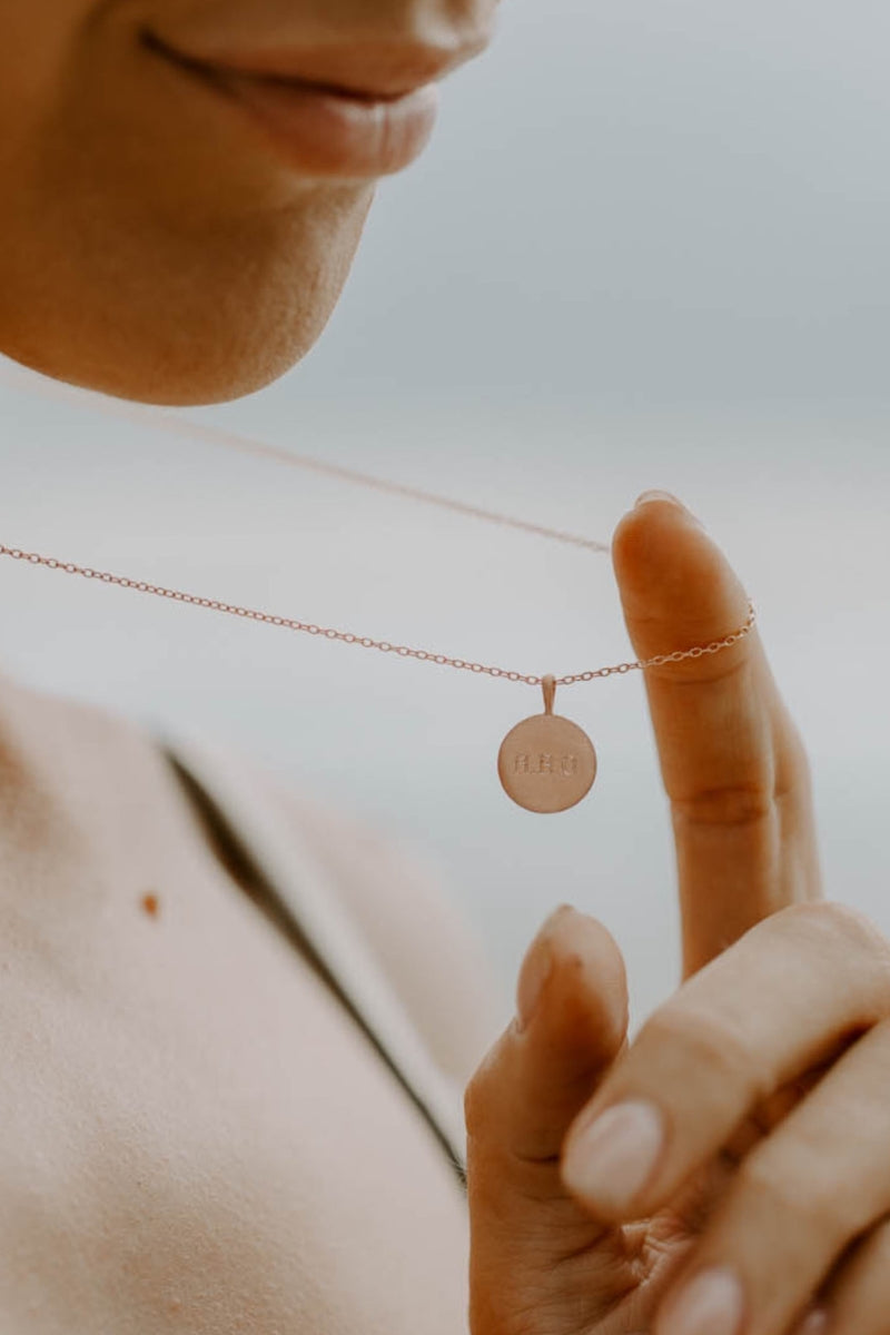 Model holding Petite Signature Necklace in rose gold by the chain to showcase the engraved round pendant in natural light.