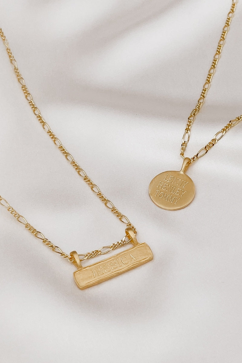Flatlay of gold Signature Necklace and Bar Necklace with engraved names on soft satin backdrop, showcasing timeless design.