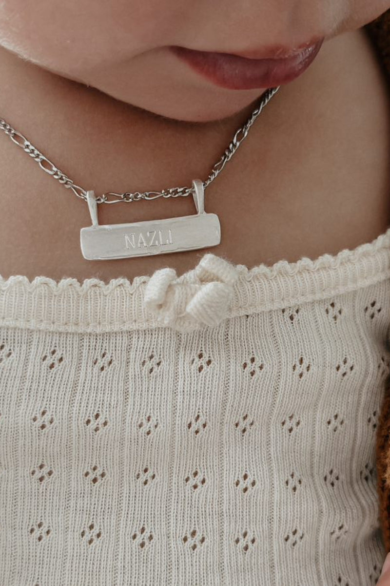 Signature Bar Necklace Silver engraved with “NAZLI” worn by a baby. A sentimental silver piece to mark milestones and memories.
