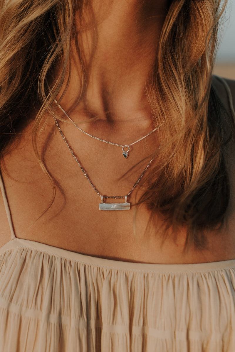 Signature Bar Necklace Silver engraved with “JOY”, layered with a fine heart charm necklace. A delicate and meaningful sterling silver piece.