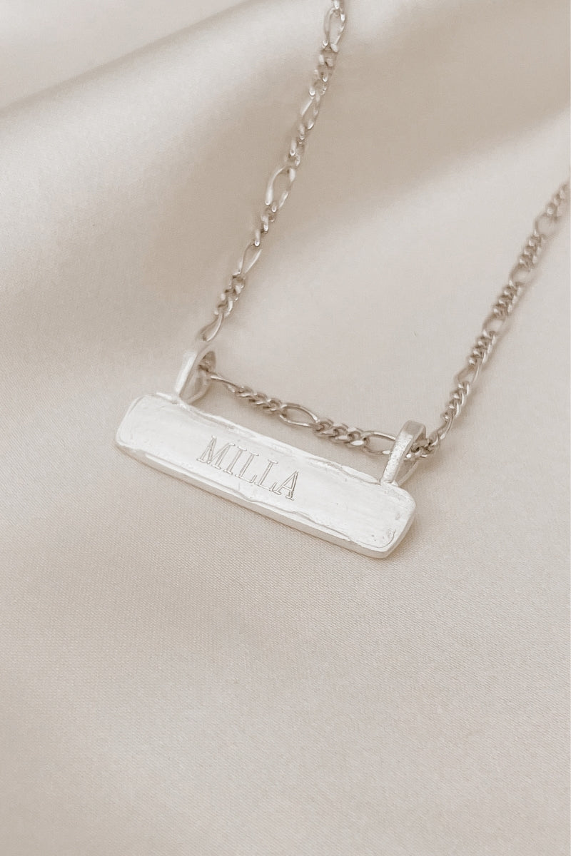 Signature Bar Necklace Silver engraved with “MILLA”, styled on soft fabric to highlight its handcrafted texture and premium sterling finish.