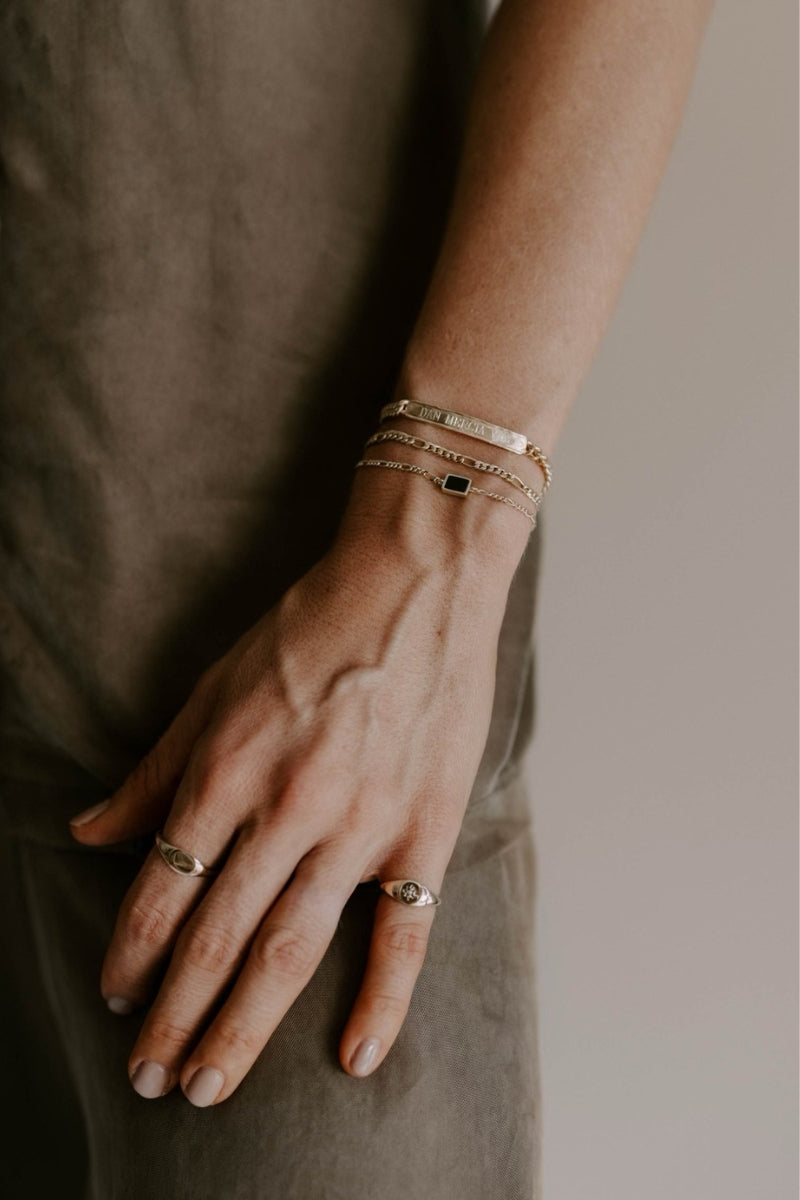 A woman wears the Signature Bracelet engraved with three names, layered with the Figaro and Loved Bracelets in gold vermeil. She also wears the personalised solid gold signet ring and Vintage Pearl Signet Ring by Bluebird Jewellery.