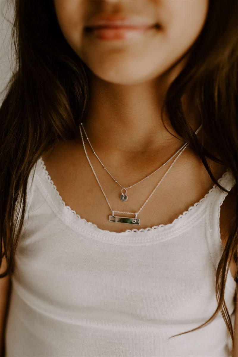 Child wearing Darling Bar Necklace in silver with heart engraving, layered with other necklaces ball chain necklace with initial engraving. 