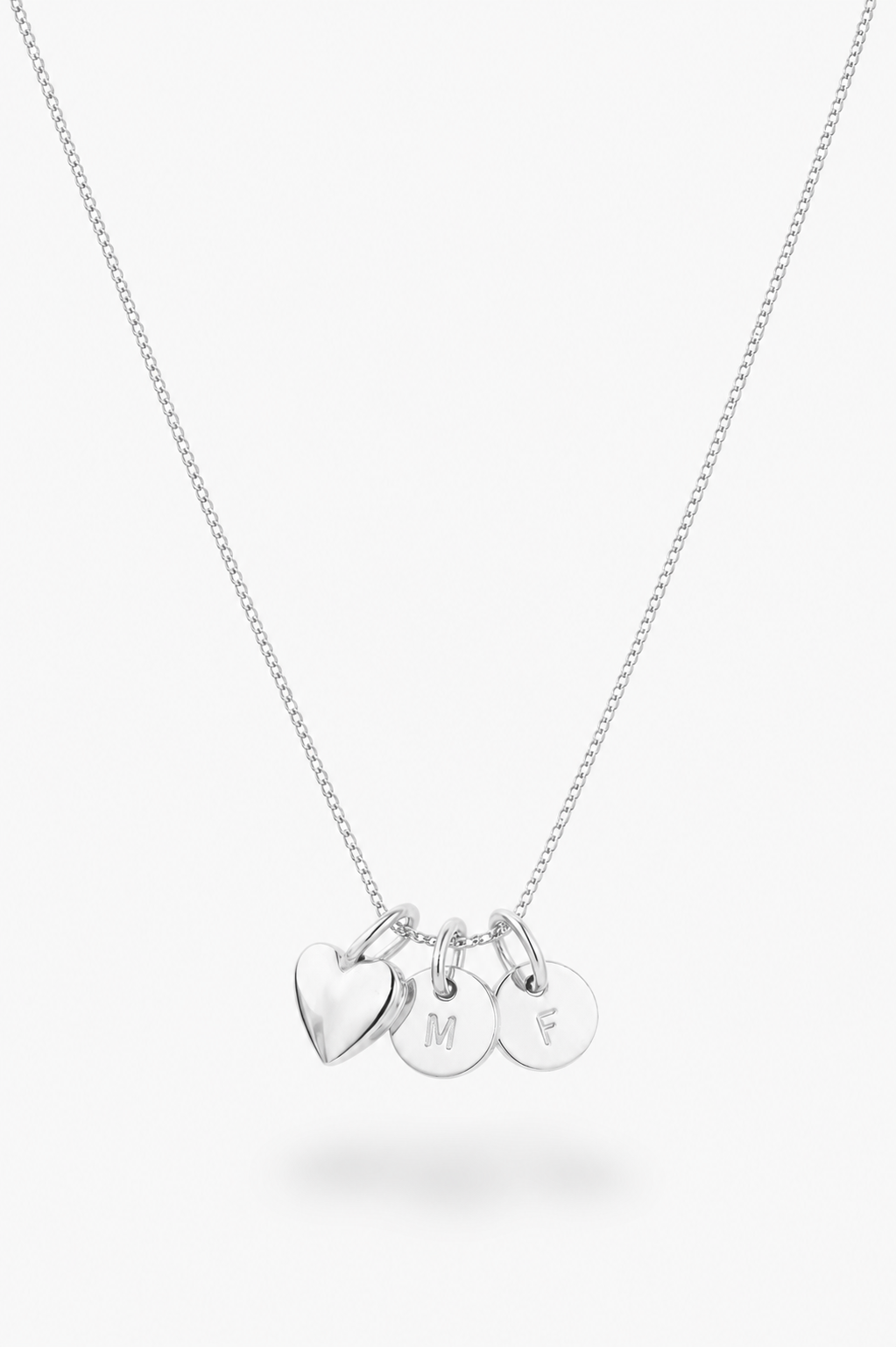 A silver chain necklace with three small hanging charms: a heart-shaped charm and two round disc charms engraved with the letters M, F, and S, shown on a light gray background.